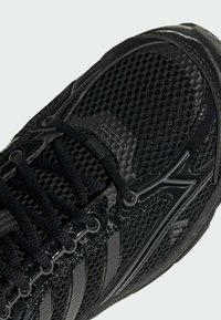 Chaussure de sport noire avec tige en mesh, surpiqûres synthétiques brillantes, lacets noirs et semelle texturée, inclinée pour montrer les détails de l'avant et du côté.