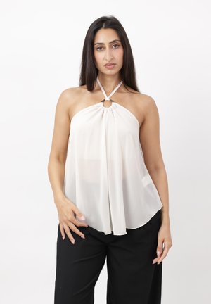 Donna con lunghi capelli scuri che indossa un top bianco senza maniche con dettaglio ad anello, abbinato a pantaloni neri larghi su uno sfondo semplice.