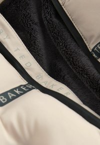 Beige Außenmaterial mit einem weichen schwarzen Pelzfutter; verfügt über "Ted Baker" Branding auf einem schwarzen Akzentstreifen und glatten Nähten.
