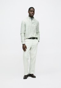 Pruhovaná zelená košeľa na zapínanie na gombíky, biele nohavice, čierny opasok, hnedé topánky. Hladká látka s jemnou textúrou a voľným strihom, oblečené na stojacom modeli.