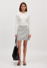 Weißes Poloshirt mit langen Ärmeln und Knopfleiste, kombiniert mit einem grauen Minirock aus Tweed mit Fransensaum. Das Model hält eine schwarze Tote-Tasche.