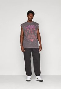 Vīrietis valkā bezpa piedurknēm tumsnējo pelēko grafisko t-kreklu, melnus bikses un baltas kedas, stāvot pret vienkāršu gaiši pelēku fonu.