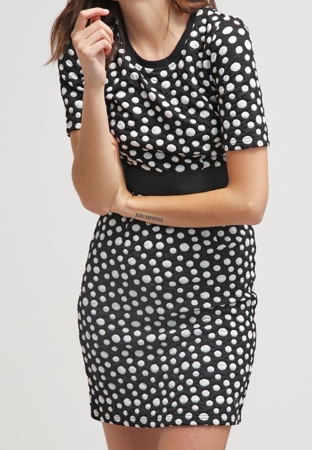 Femme portant une robe noire à manches courtes avec des pois blancs, les bras croisés, une main près du visage, montrant un tatouage de chiffre romain sur son avant-bras.