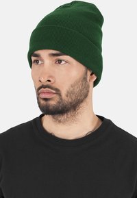 Căciulă tricotată verde cu o textură late. Se potrivește strâns pe cap, punând în evidență un design simplu, fără ornamentații, pe un cămașă neagră.