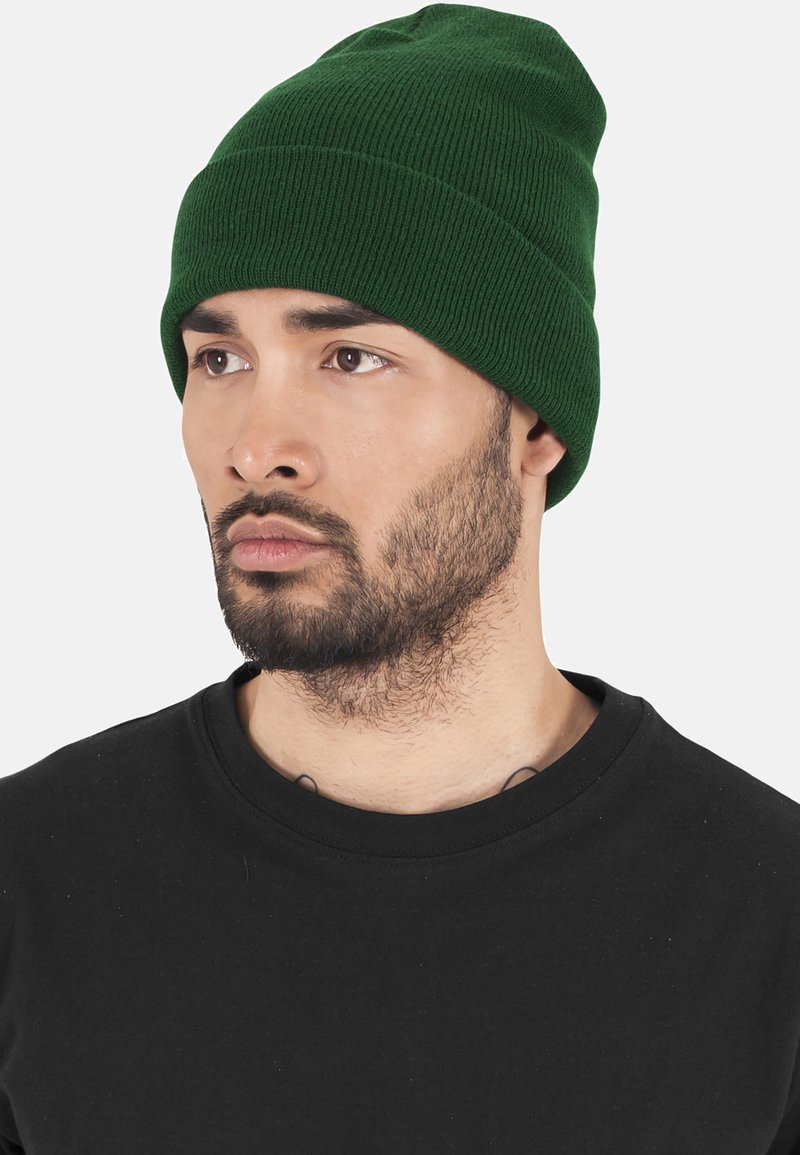 Căciulă tricotată verde cu o textură late. Se potrivește strâns pe cap, punând în evidență un design simplu, fără ornamentații, pe un cămașă neagră.