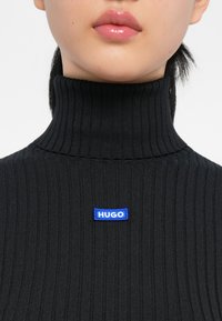 Maglione nero a coste con collo alto e tag HUGO blu al centro. Il tessuto appare testurizzato e aderente.