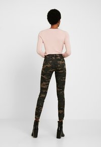 Jean camouflage au design ajusté dans des teintes de vert et de marron, associé à un haut à manches longues rose clair et des bottes à talons hauts noires.