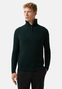 Blonder Mann in einem dunkelgrünen, gerippten Pullover mit halbem Reißverschluss und schwarzer Hose, steht mit einer Hand in der Tasche vor einem schlichten Hintergrund.