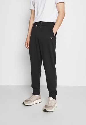 Mann trägt schwarze Jogginghose mit Kordelzug in der Taille, weißes T-Shirt und beige Sneaker, steht vor einer schlichten hellen Wand.