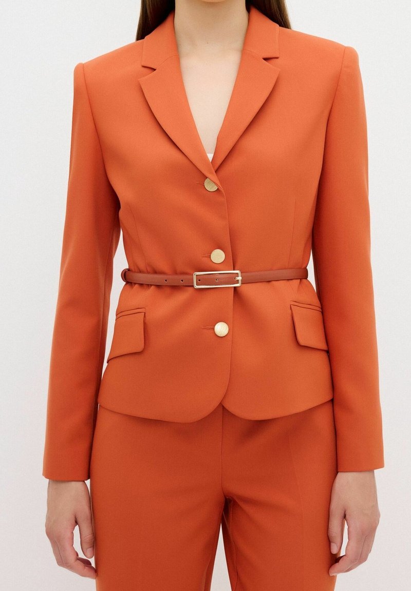 Femme portant un blazer orange cintré avec des boutons dorés et une fine ceinture marron, assorti à un pantalon orange.