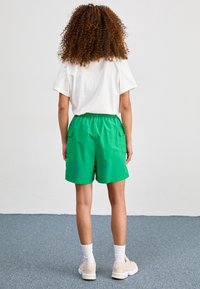 Pantalones cortos verdes con cintura elástica y bolsillos laterales, hechos de tela ligera. Combinados con una camiseta blanca y zapatillas crema.