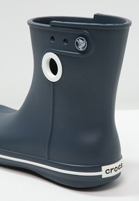 Botte en caoutchouc bleu foncé avec une rayure blanche à la base, un trou de ventilation rond et un logo circulaire sur le côté. Texture lisse.