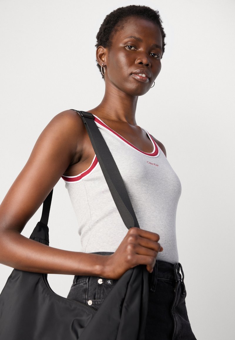 Hellgraues Tanktop mit rotem Saum und besticktem Logo; das Modell hält eine schwarze Schultertasche und trägt dunkle Jeans.