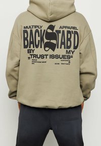 Sudadera color caqui con gran texto negro en la parte posterior que dice "TRAICIONADO POR MIS PROBLEMAS DE CONFIANZA." Material suave, mangas largas y una capucha amplia.