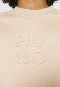 Sudadera beige claro con un logotipo "HUGO" texturizado y en relieve en el pecho, con un cuello redondo acanalado y una tela suave.