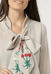 Blusa beige con cuello atado; presenta un colorido bordado floral en rojo, blanco y verde. Tejido suave con una textura y caída lisas.