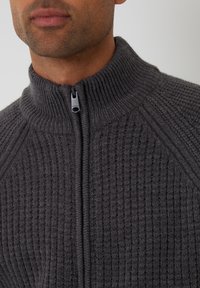 Suéter gris con cremallera, de canalé y cuello alto. Textura suave y costuras en contraste. La cremallera tiene un tirador negro.