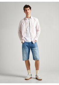Licht gestreepte roze langemouwen shirt over een wit graphic T-shirt, gecombineerd met blauwe denim shorts en witte sneakers met crèmekleurige accenten.