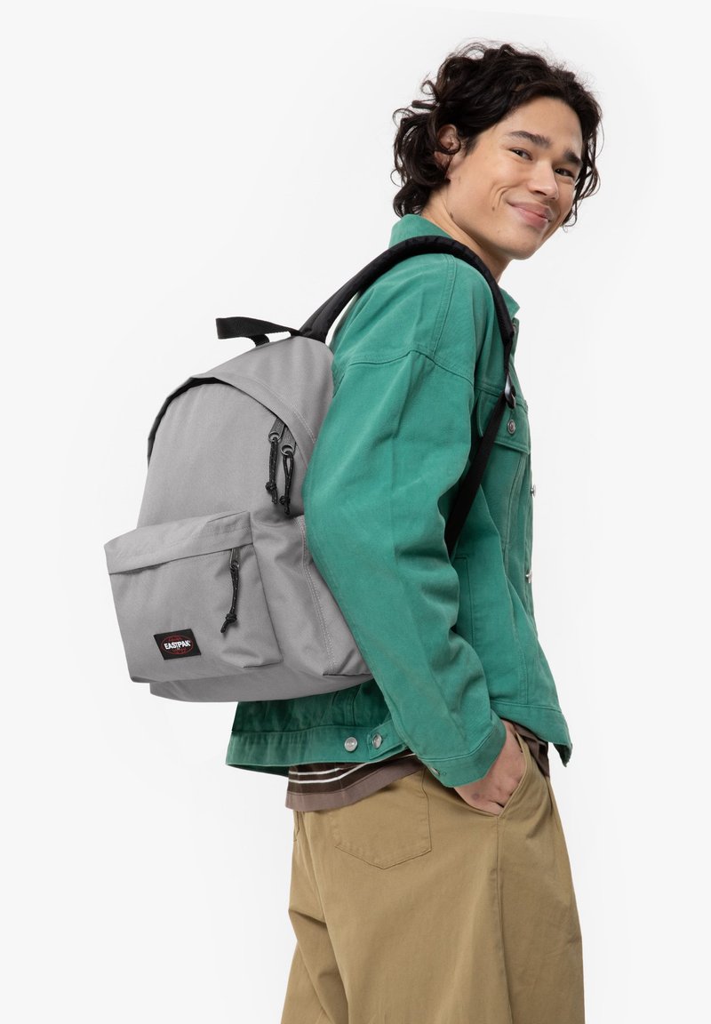 Mochila gris que presenta un compartimento principal, un bolsillo frontal, correas ajustables y herrajes negros, llevada por una persona con una chaqueta verde.