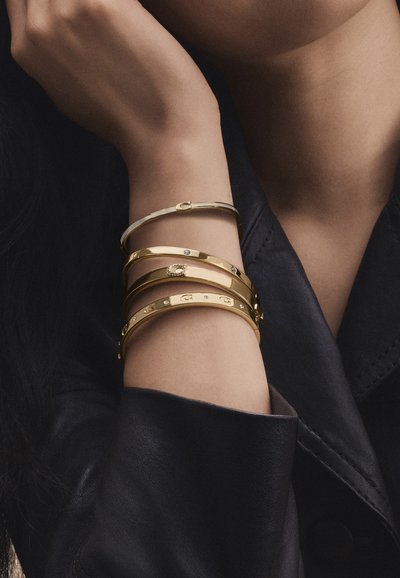 Bras portant quatre bracelets en or avec de petits détails décoratifs sur un fond de veste en cuir foncé.