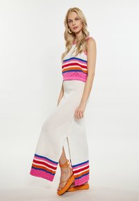 Gebreide jurk met een witte basis, meerkleurig gestreept patroon op het bovenlichaam en zijsplitten. Gepaard met oranje sandalen.