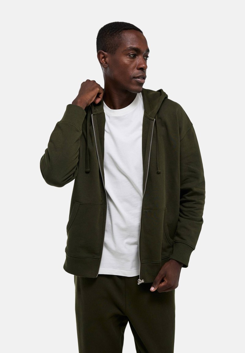 Homme portant un sweat à capuche zippé olive et un pantalon avec un t-shirt blanc, regardant sur le côté avec une main ajustant la capuche.