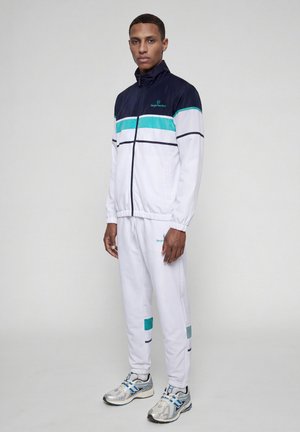 Joven de pie, vistiendo un chándal blanco con rayas azul marino y turquesa y zapatillas de correr plateadas sobre un fondo claro y sencillo.