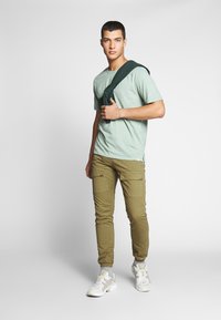 Grønn kortermet skjorte, khaki cargo-bukser med lommer og hvite joggesko med grå detaljer, poserer mot en ensfarget bakgrunn.