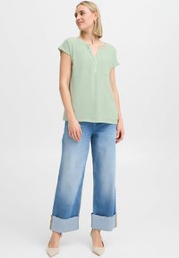 Helles grünes Kurzarmblusenshirt mit V-Ausschnitt und aufgesetzter Tasche, kombiniert mit weiten hellblauen Jeans mit umgeschlagenen Säumen und fransigen Kanten.