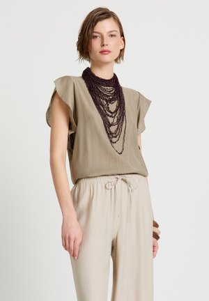 Donna con pantaloni beige a gamba larga, camicetta verde oliva a maniche corte, collana a strati con perline viola scuro e braccialetti di legno su sfondo bianco.