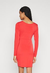 Robe rouge à manches longues avec une silhouette ajustée, dos plongeant et texture de tissu lisse, se terminant au-dessus du genou.