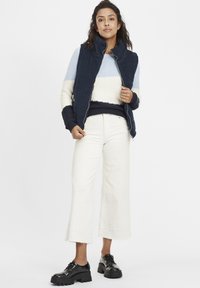 Marinblå quiltad väst över en ljusrandig tröja, i kombination med vita vida jeans och svarta platåskor. V-ringad design med ribbad textur.