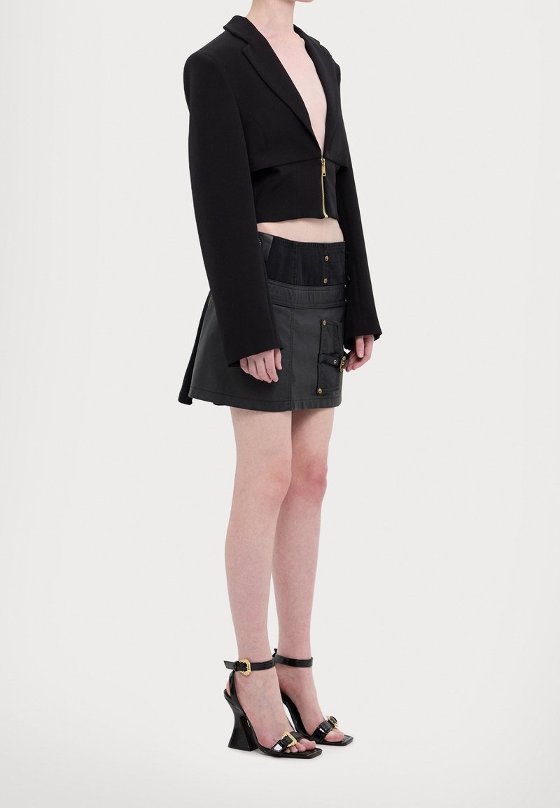 Svart kort blazer med långa ärmar och gulddragkedjedetalj, i kombination med en högmidjad svart minikjol med fickor och guldaccenter.