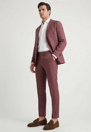 Man standing wearing a tailored mauve suit with a white shirt and brown tassel loafers against a plain light background.