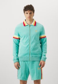 Ensemble de survêtement en velours bleu clair avec une veste zippée. Comprend un col et des accents de manches rayés rouges, jaunes et blancs. Détail du logo en or.