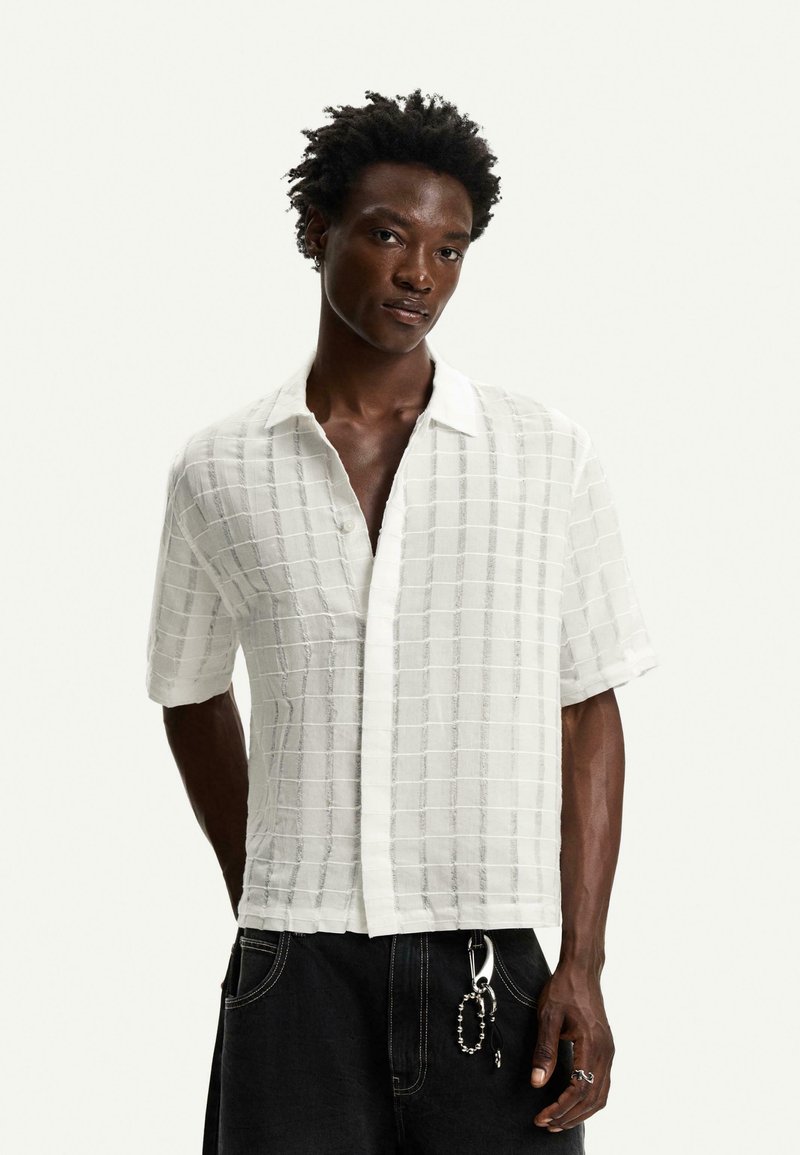 Camicetta bianca a maniche corte con texture a quadri, colletto e cierre con bottoni. Indossata con pantaloni neri e accessori in catena argentati.