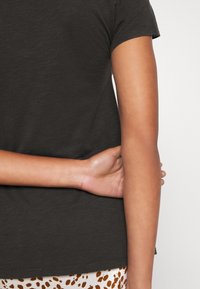 Eine Person trägt ein dunkelgraues Hemd und einen gemusterten Rock, während sie mit verborgenen Händen hinter dem Rücken vor einem schlichten hellen Hintergrund steht.