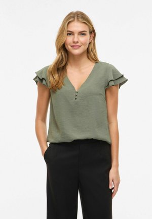 Blondynka w teksturowanej oliwkowozielonej bluzce z falbaniastymi rękawami i czarnych spodniach, stojąca z jedną ręką w kieszeni.