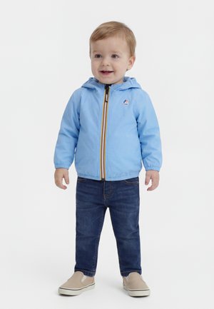 Batole sa usmieva, má na sebe svetlomodrú bundu s kapucňou, tmavomodré rifle a béžové tenisky bez šnúrok, stojí na bielom pozadí.