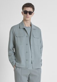 Giacca azzurro chiaro, strutturata, con due tasche sul petto, chiusura con bottoni e colletto. Indossata sopra una camicia coordinata dello stesso colore.