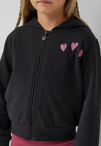 Zwarte zip-up hoodie van zacht materiaal, met een aansluitende tailleband, manchetten aan de mouwen en drie glinsterende roze hartaccenten op de borst.