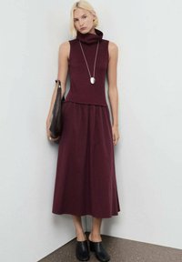 Una mujer con cabello corto rubio lleva un vestido sin mangas burdeos, un largo collar de plata, carga un bolso marrón oscuro y unos mule negros con tacón.