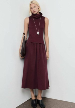 Femme aux cheveux blonds courts portant une robe sans manches bordeaux, un long collier en argent, tenant un sac marron foncé et des mules noires à talons.