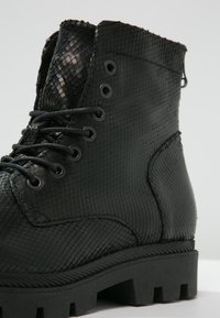 Czarny botek z ekologicznej skóry z teksturą przypominającą skórę gada, z przodem na sznurowadła i masywną gumową podeszwą. Posiada metalowe przelotki i zamek błyskawiczny.