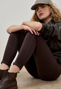Jeans marrones ajustados, chaqueta de cuero negra y una gorra de cuadros beige. El modelo está sentado con las piernas cruzadas, con botas tobilleras marrones en el suelo.