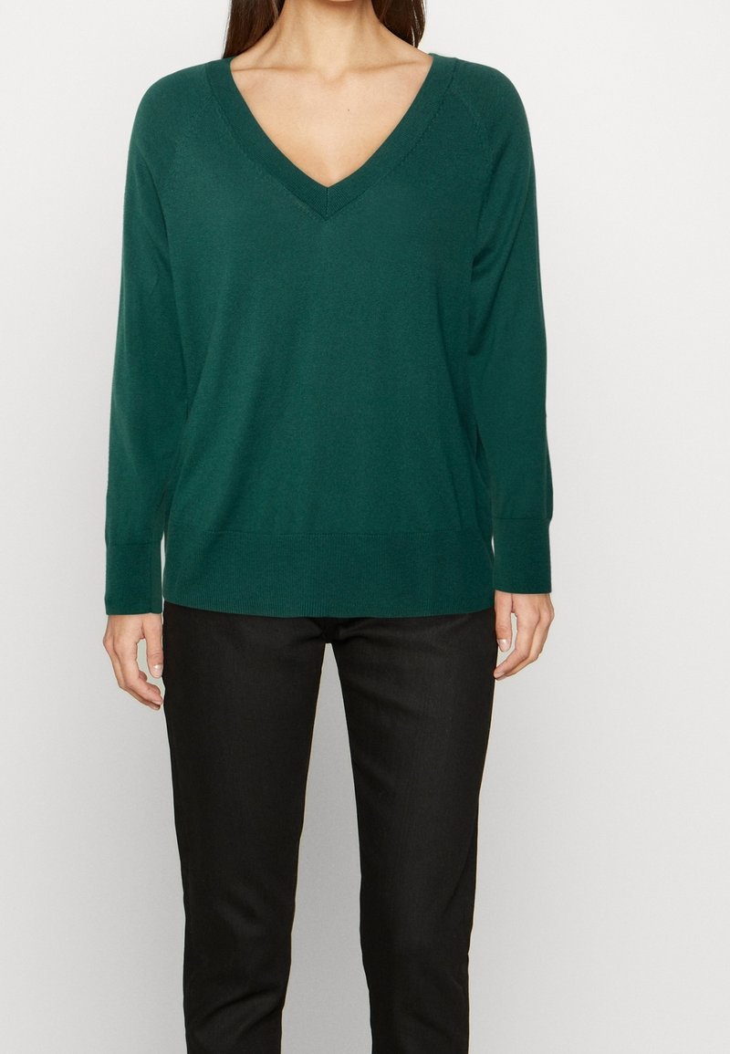 Femme portant un pull à manches longues col en V vert foncé et un pantalon noir, debout devant un fond clair uni.