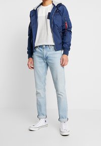 Marineblå bomberjacka med grå huva, ficka med dragkedja och elastiska ärmslut; tillsammans med randig t-shirt och ljusblå jeans; vita sneakers.