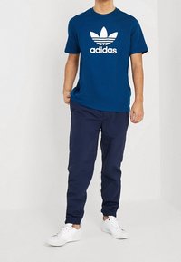 Modrá bavlněná tričko s bílým logem Adidas a grafikou trojlístku, spárováno s tmavě modrými kalhotami a bílými teniskami. Volný střih a hladká textura.