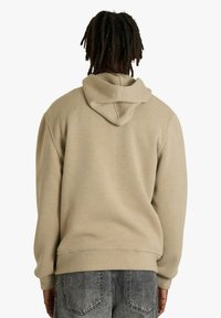 Felpa beige con cappuccio in tessuto morbido, caratterizzata da una vestibilità ampia e polsini a coste, vista da dietro. Jeans grigi visibili sotto.