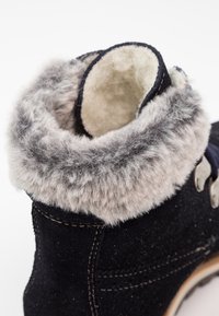 Schwarze Wildlederstiefel mit einem weichen grauen Kunstfellkragen, gestickten Details und metallischen Hardware-Akzenten. Glattes Innenfutter sichtbar.